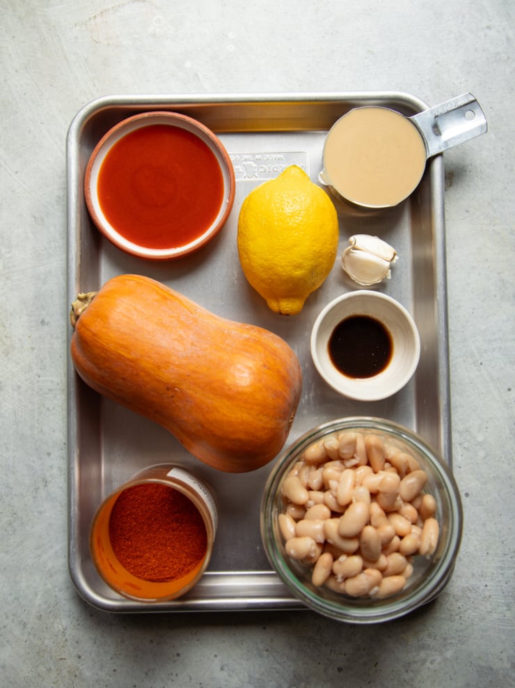 Ingredients for butternut squash dip on a sheet pan: a whole honeynut squash, buffalo hot sauce, lemon, tahini, garlic, vegan Worcestershire, paprika, and white beans.