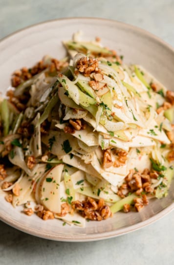 Close-up of celery root apple salad in a cream bowl, with shaved celery root, fennel, apple, and celery tossed with parsley and topped with maple candied walnuts.