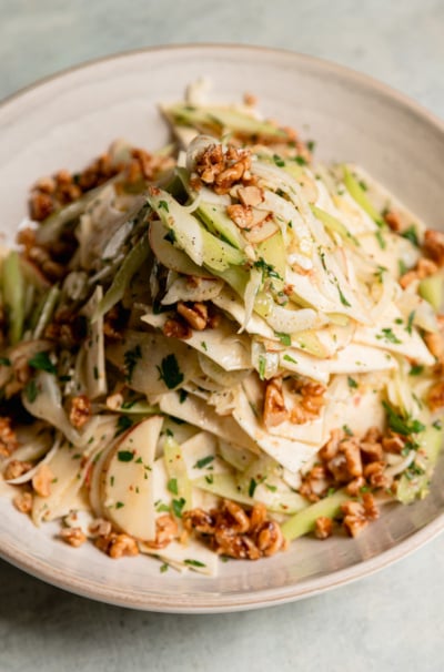 Close-up of celery root apple salad in a cream bowl, with shaved celery root, fennel, apple, and celery tossed with parsley and topped with maple candied walnuts.