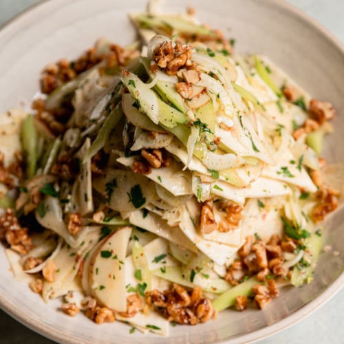 Close-up of celery root apple salad in a cream bowl, with shaved celery root, fennel, apple, and celery tossed with parsley and topped with maple candied walnuts.