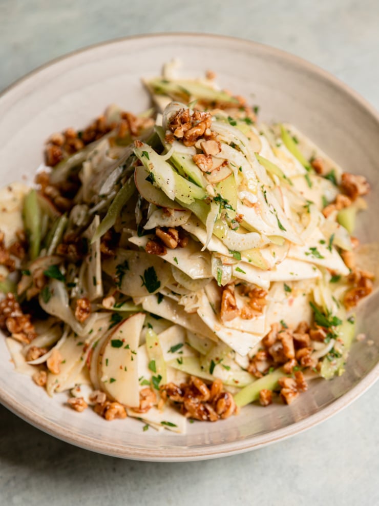 Close-up of celery root apple salad in a cream bowl, with shaved celery root, fennel, apple, and celery tossed with parsley and topped with maple candied walnuts.