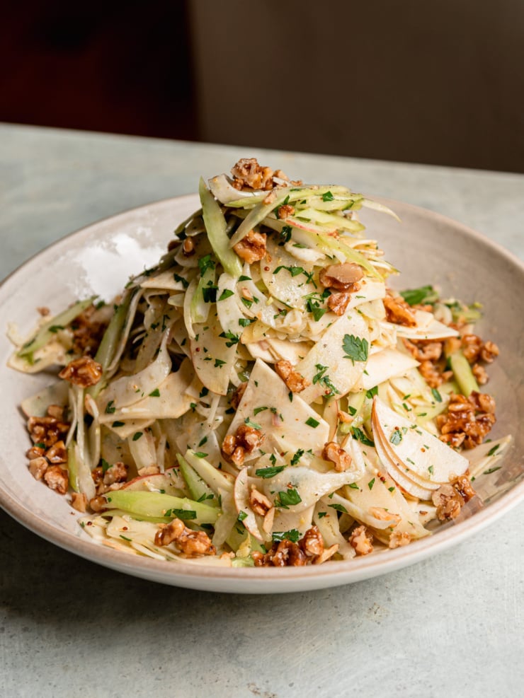 A tall pile of celery root apple salad in a shallow bowl, with candied walnuts scattered around the edges.