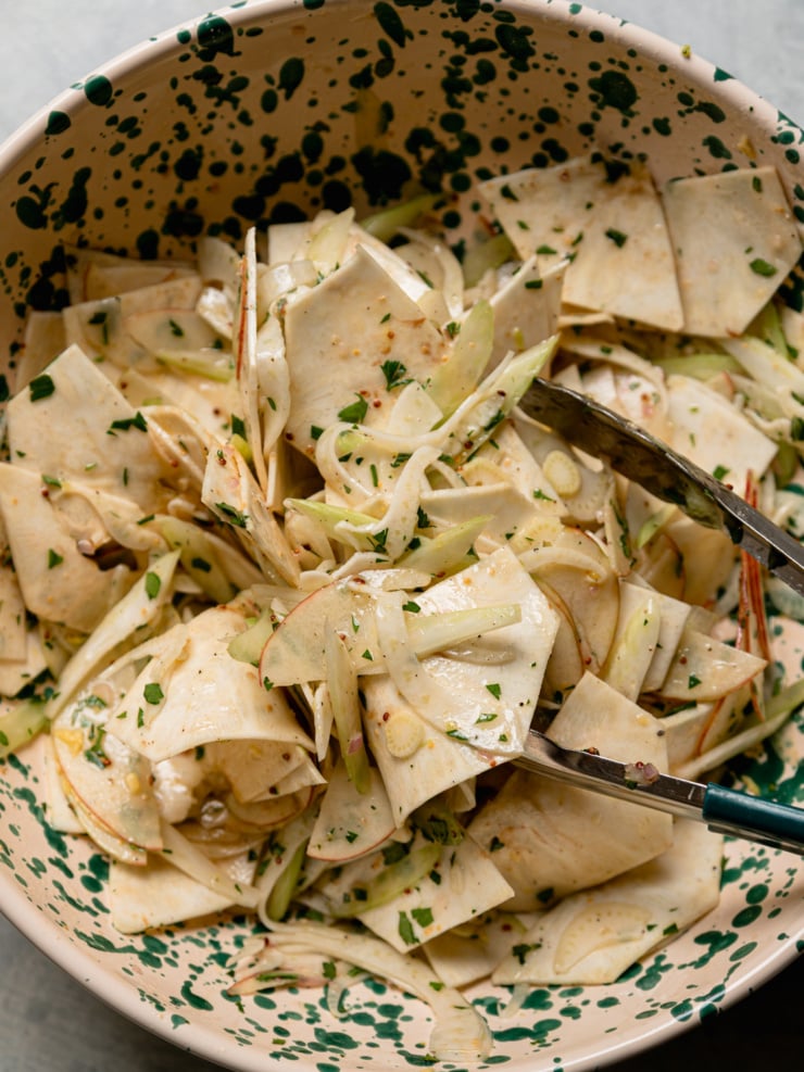 Tongs toss shaved celery root, fennel, apple, and celery in a sherry maple vinaigrette in a green speckled bowl, for a celery root apple salad.