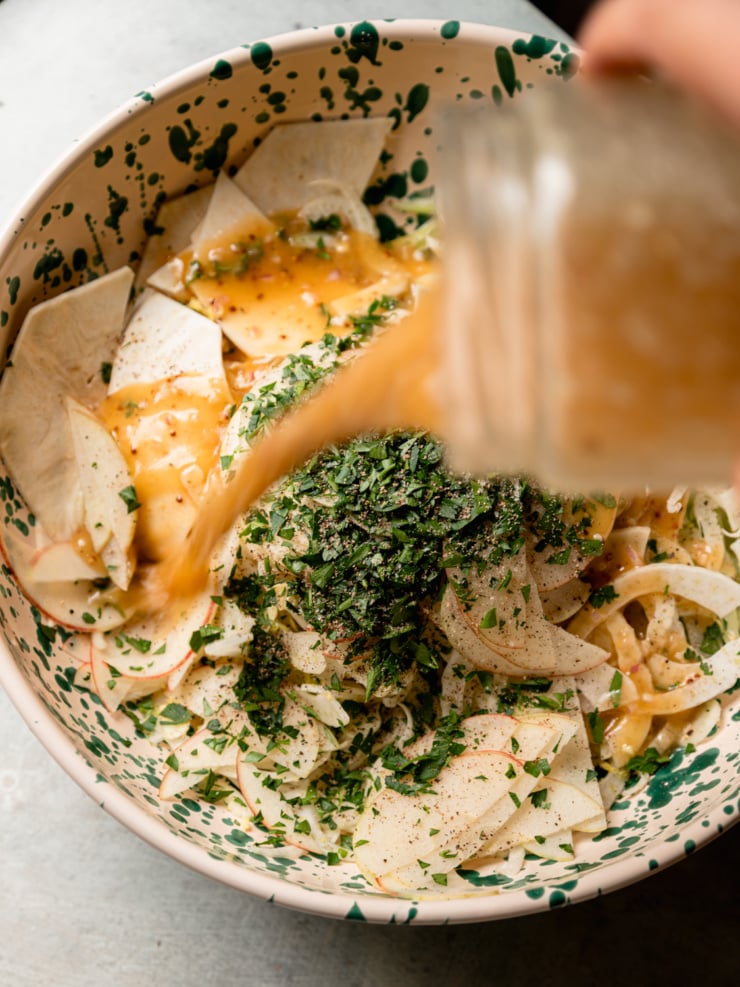 Sherry maple vinaigrette pours from a jar over shaved vegetables, chopped parsley, and black pepper in a speckled bowl.