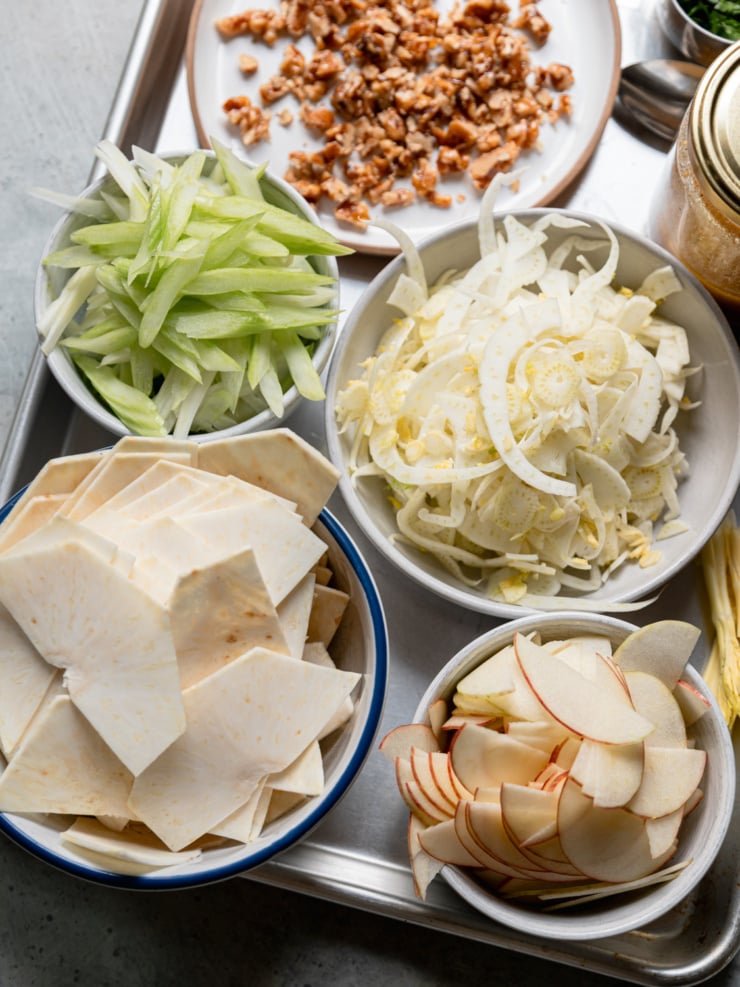 Bowls of shaved celery root, fennel, apple slices, and thinly sliced celery on a sheet pan, with a plate of maple candied walnuts and a jar of vinaigrette.