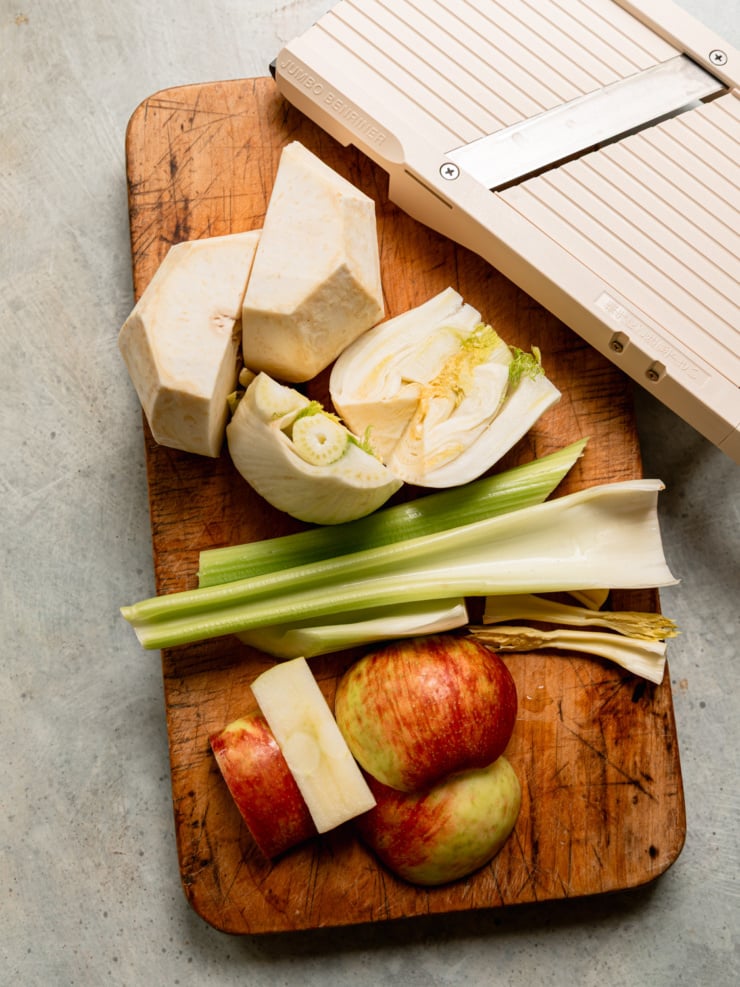 Peeled celery root, a halved fennel bulb, celery stalks, and a cut apple on a wooden cutting board next to a mandoline.