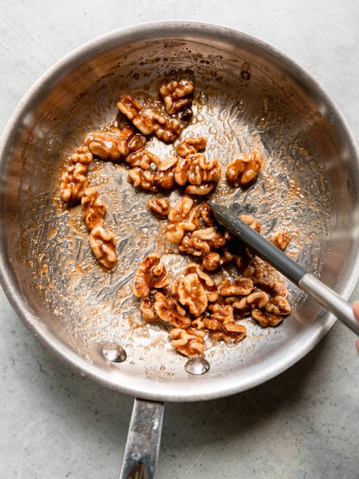 Walnut halves bubbling in maple syrup in a stainless steel skillet as they’re stirred.