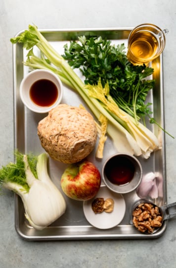 Ingredients for celery root apple salad on a sheet pan: a whole celery root, fennel bulb, apple, celery, parsley, shallots, walnuts, olive oil, sherry vinegar, maple syrup, and two mustards.