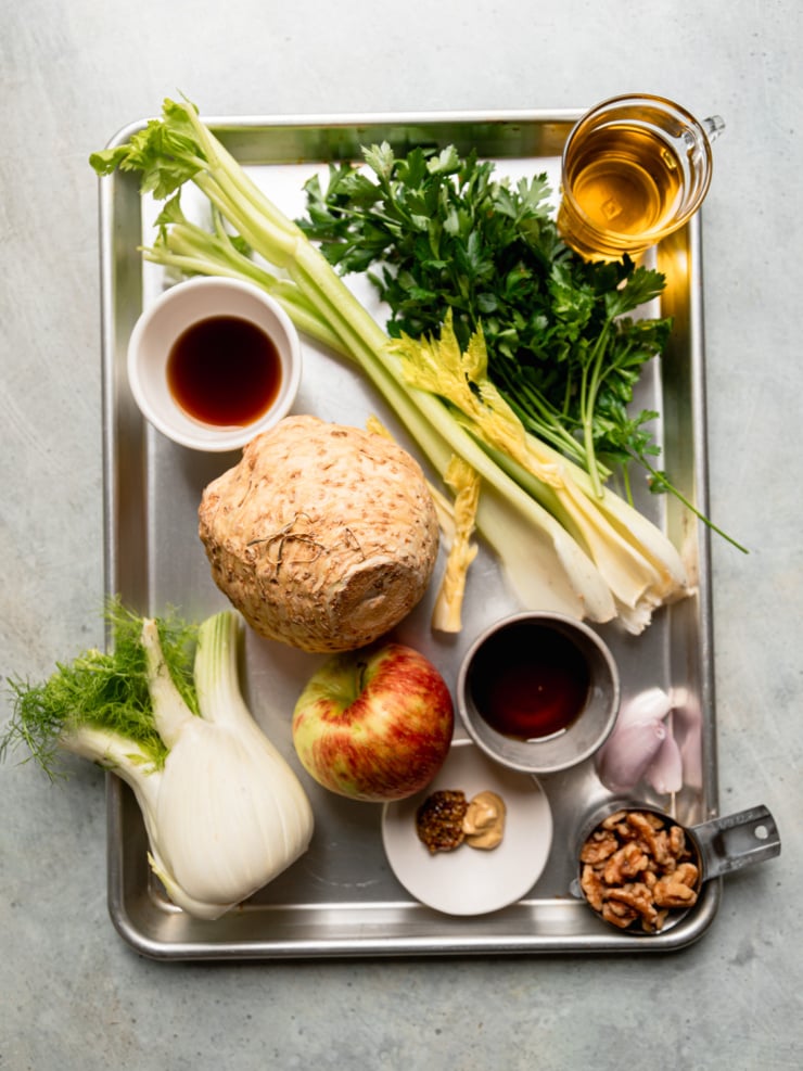 Ingredients for celery root apple salad on a sheet pan: a whole celery root, fennel bulb, apple, celery, parsley, shallots, walnuts, olive oil, sherry vinegar, maple syrup, and two mustards.