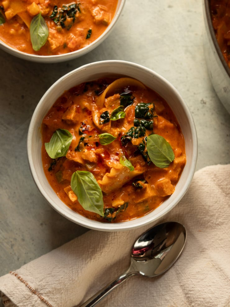 Vegan lasagna soup in a white bowl topped with fresh basil and chili oil, with a spoon and linen napkin alongside.