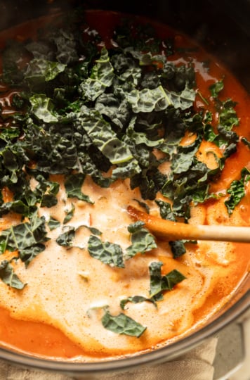 Chopped lacinato kale and savory cashew cream being stirred into tomato broth with a wooden spoon.