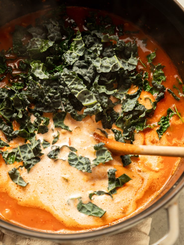 Chopped lacinato kale and savory cashew cream being stirred into tomato broth with a wooden spoon.