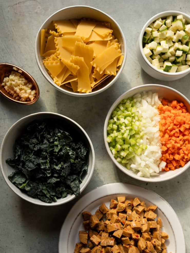 Prepped ingredients for vegan lasagna soup: broken lasagna noodles, diced zucchini, garlic, kale, mirepoix, and chopped veggie sausage.