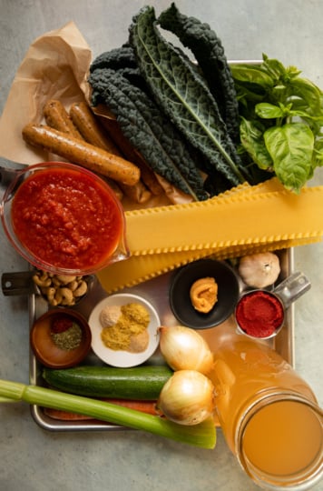 Veggie sausages, kale, basil, marinara, lasagna noodles, cashews, spices, miso, tomato paste, onions, zucchini, celery, carrot, and vegetable stock on a tray.