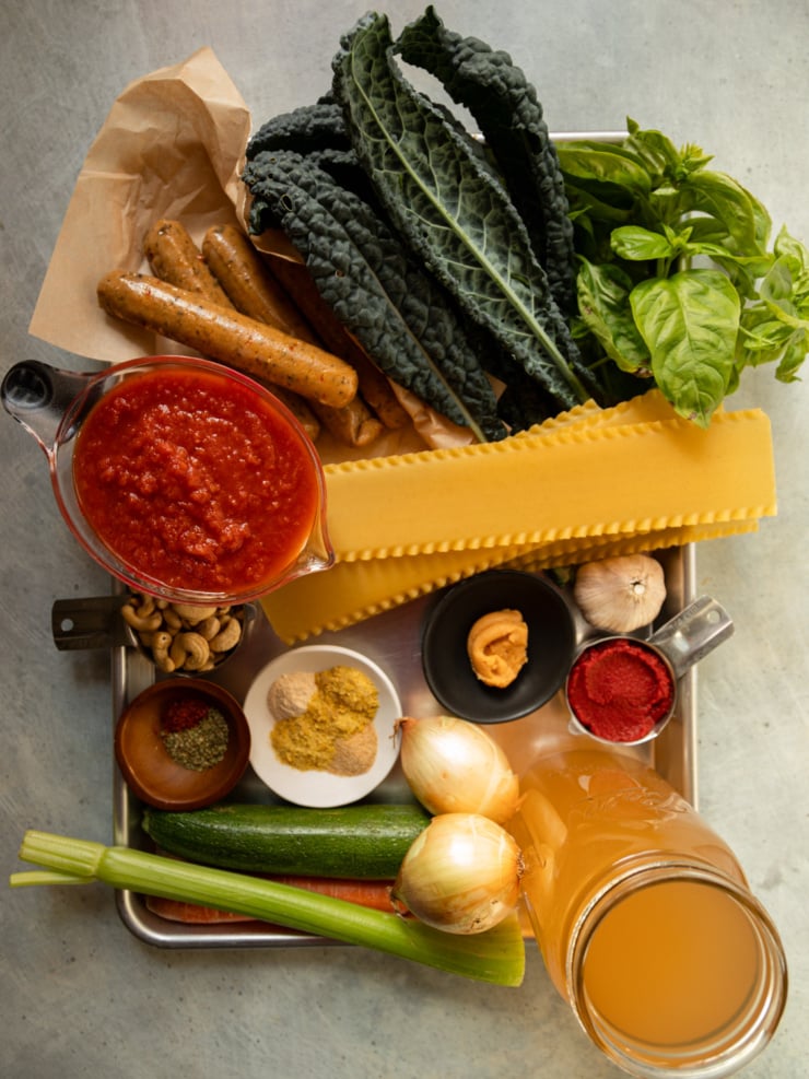 Veggie sausages, kale, basil, marinara, lasagna noodles, cashews, spices, miso, tomato paste, onions, zucchini, celery, carrot, and vegetable stock on a tray.