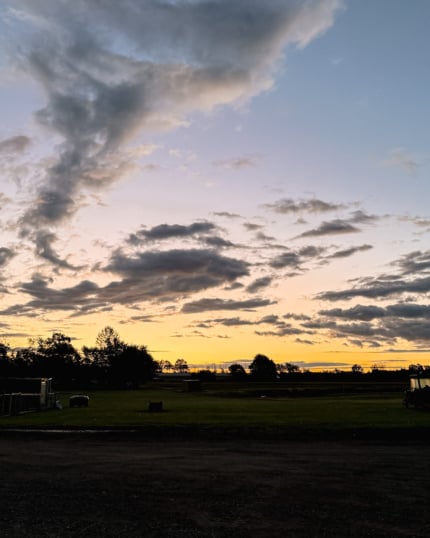 A sunset over a farm field, with dark clouds drifting across a blue and golden sky and trees silhouetted along the horizon.