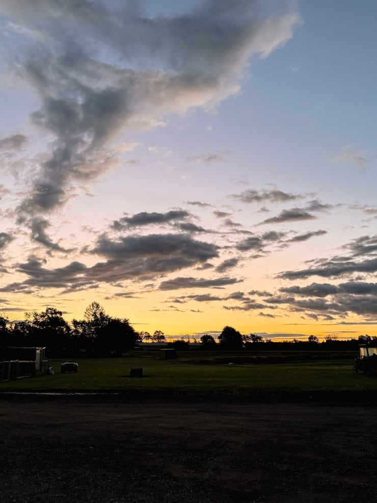 A sunset over a farm field, with dark clouds drifting across a blue and golden sky and trees silhouetted along the horizon.