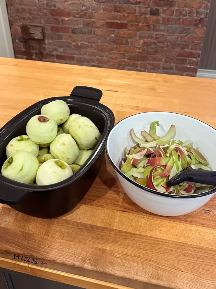 A black pot full of peeled apples sits next to a white enamel bowl of red and green apple peels and a peeler on a butcher block counter, with a brick wall behind.